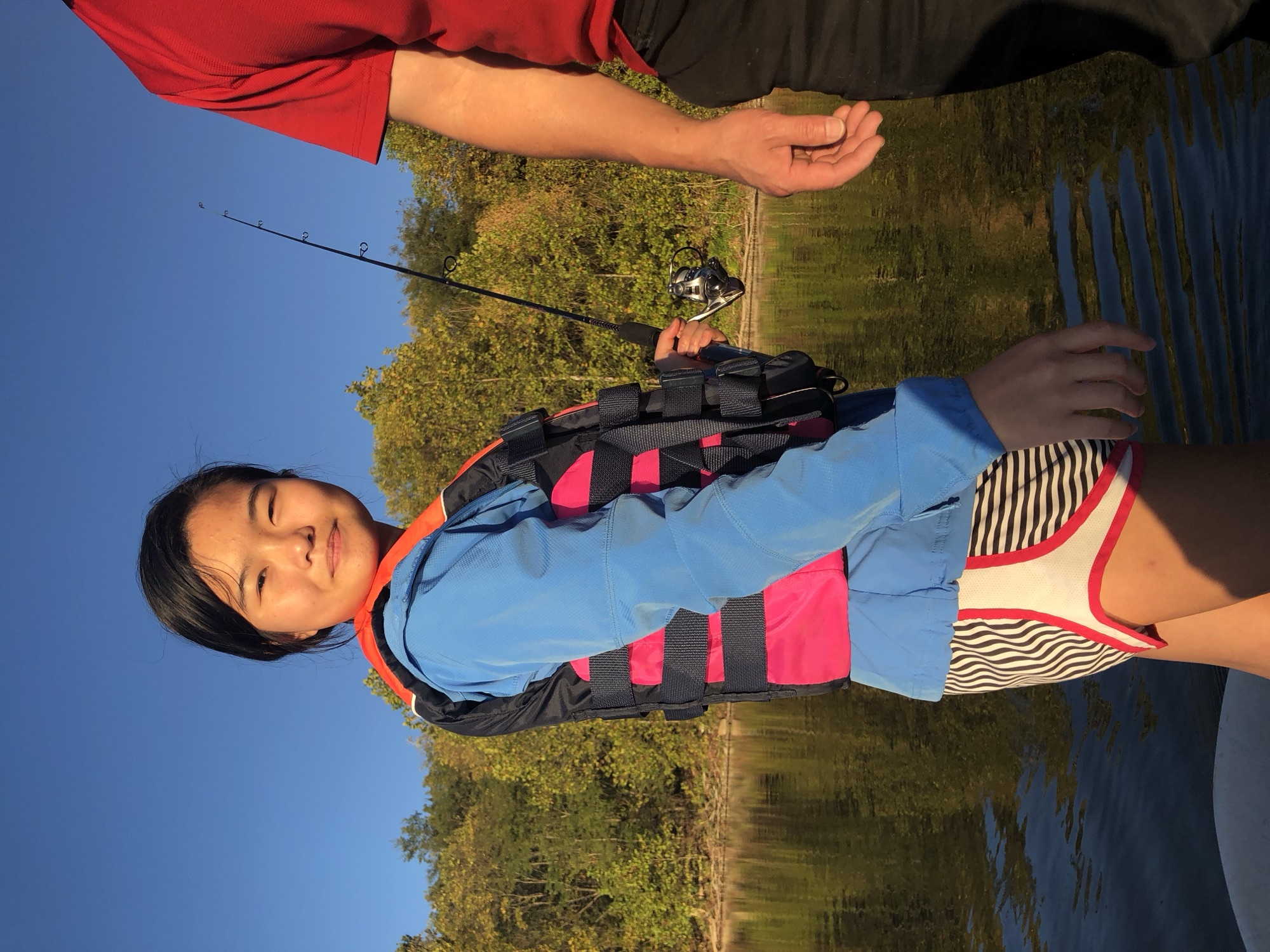 Jade Zhao standing in a small boat on a still lake, holding a fishing rod. She wears a blue jacket and a pink and black life jacket; trees and a clear sky reflect in the water.