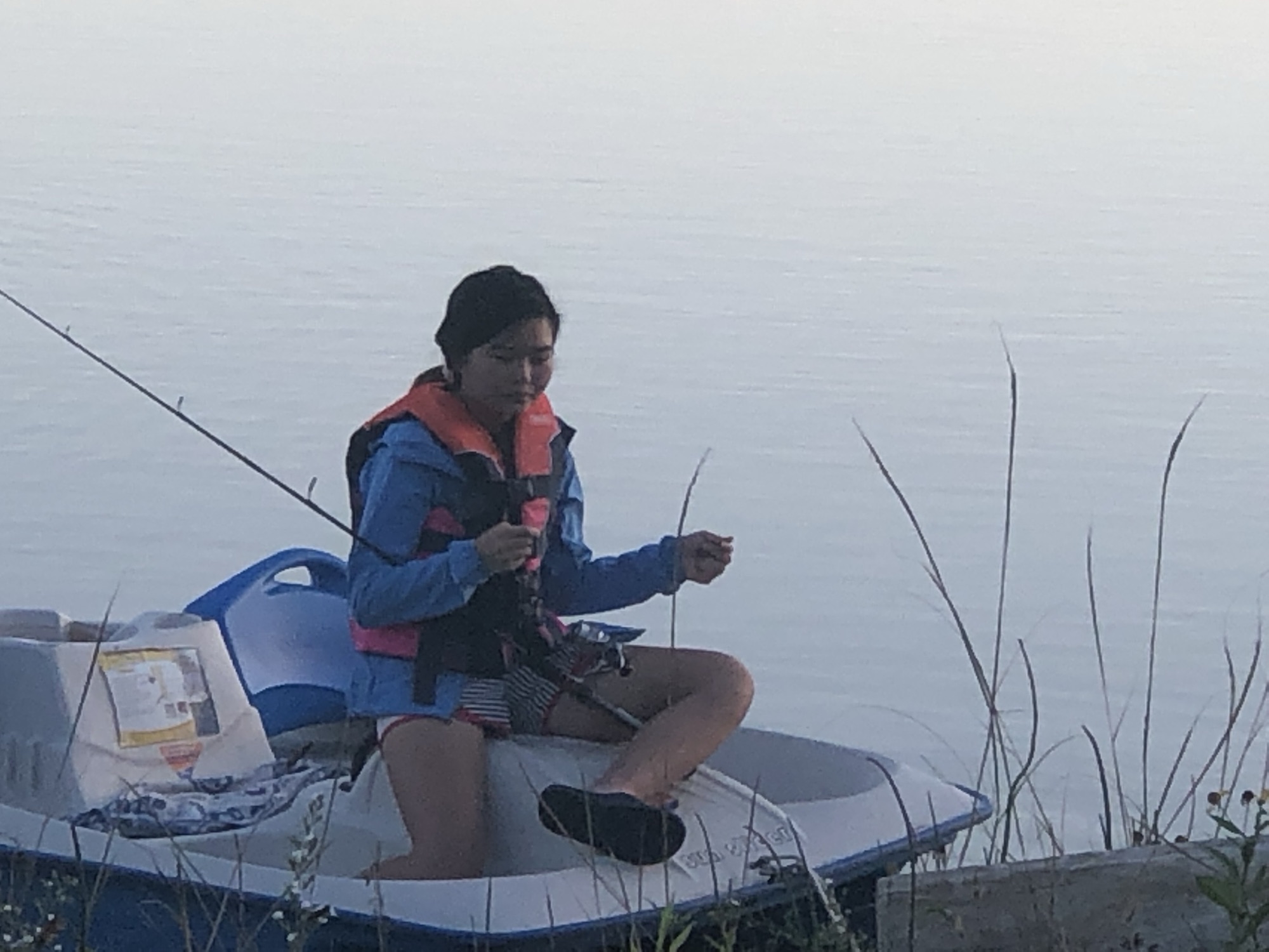 Jade Zhao sitting cross-legged in a small white and blue boat near a grassy shore, looking down at a fishing rod. She wears a blue jacket and an orange life vest; pale water stretches behind her.