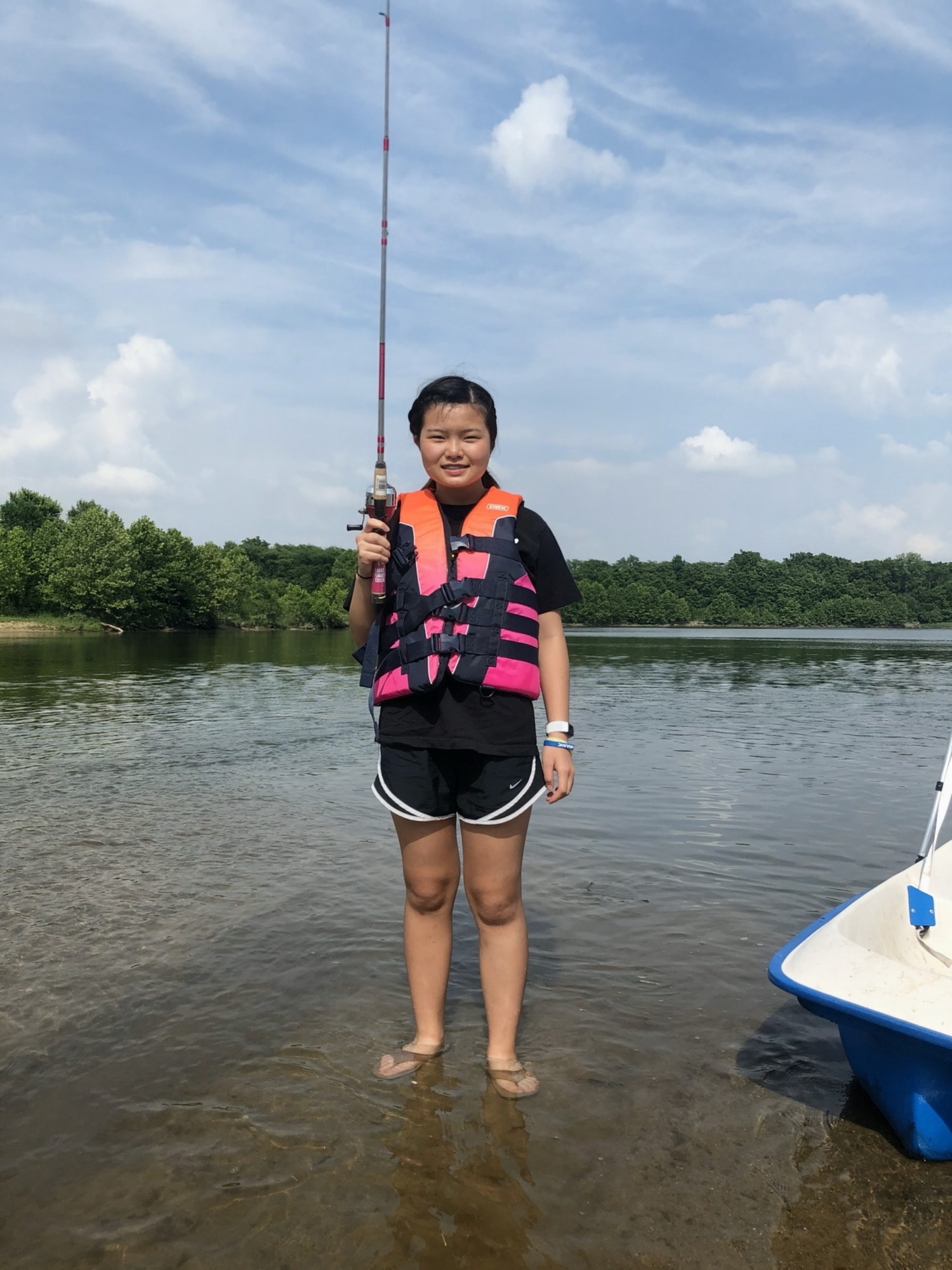Jade Zhao standing in shallow water at the edge of a lake, holding a fishing rod upright. She wears a pink and navy life jacket; a white boat sits to the right and a treeline meets a blue sky.
