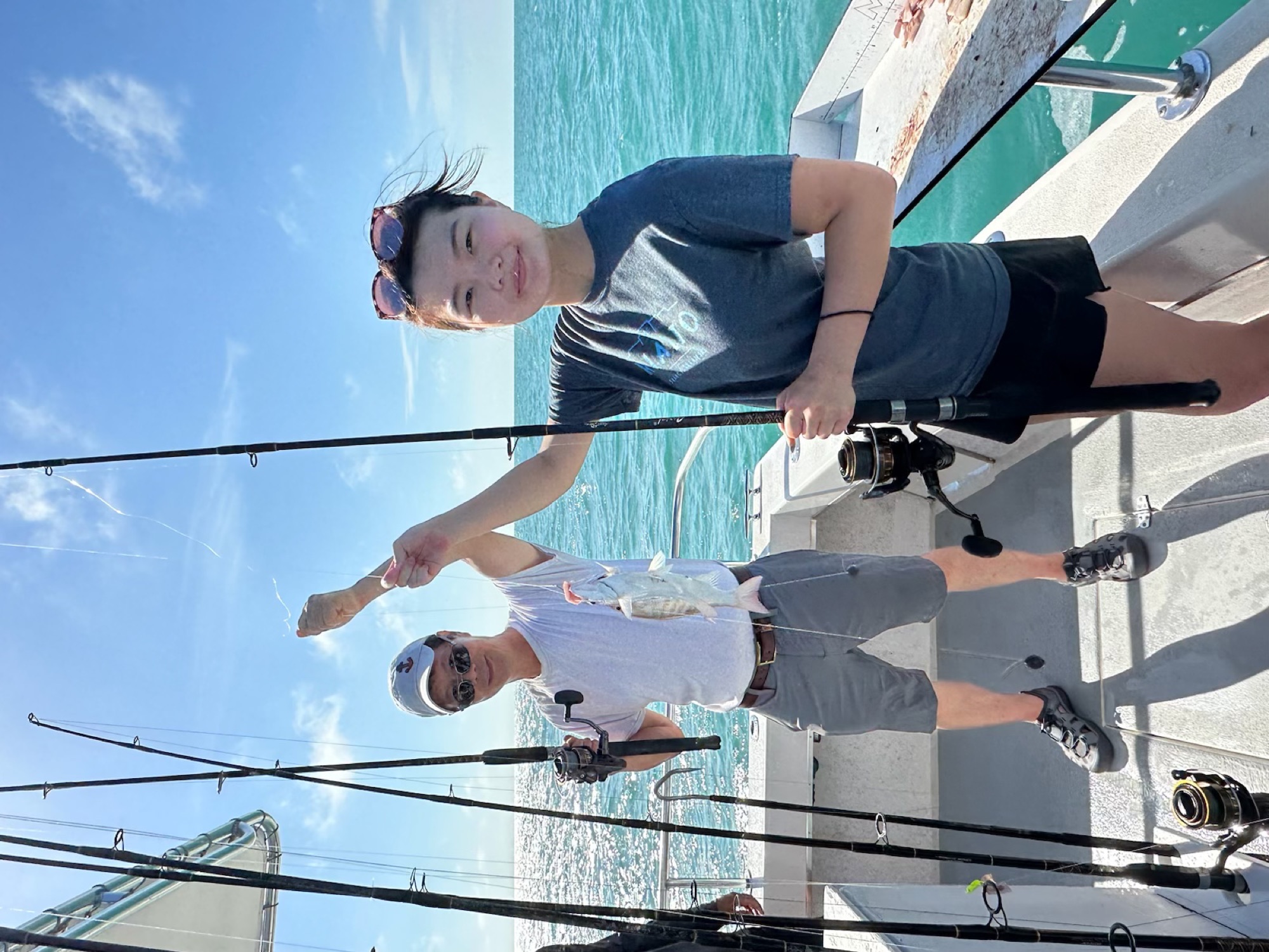 Jade Zhao on a boat at sea holding a fishing rod with a small silver fish on the line, standing beside an older companion who also holds the line. Turquoise water, extra rods in holders, and a bright sky.