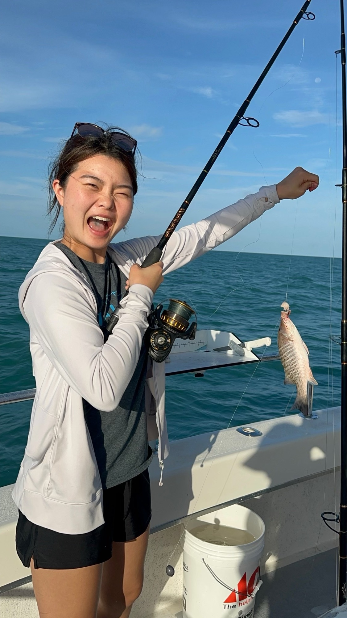 Jade Zhao on a boat at sea, cheering with her mouth open, holding a fishing rod high while a small silvery fish hangs from the line. Grey hoodie, sunglasses on her head, turquoise water and a clear sky behind the deck.