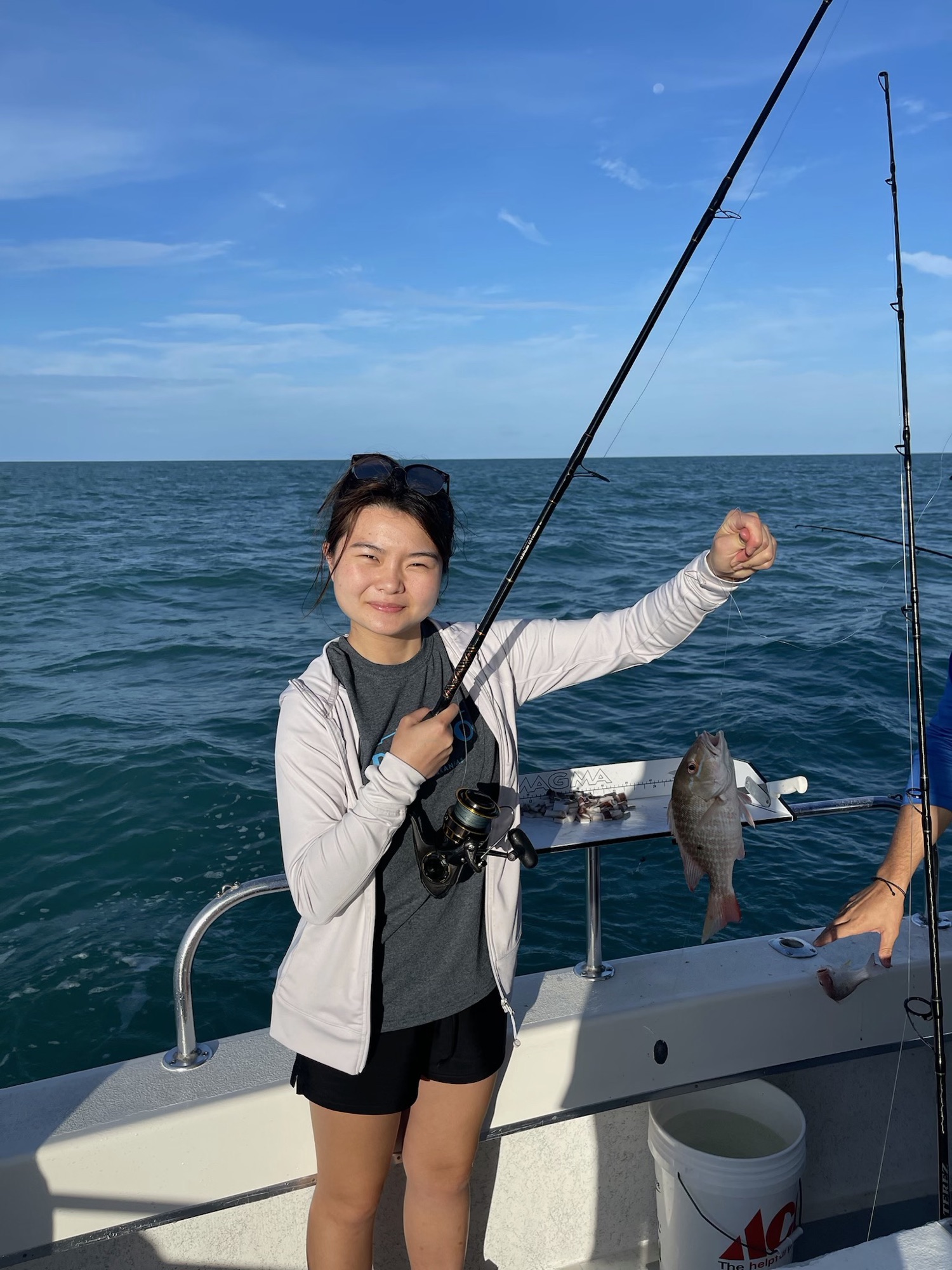 Jade Zhao standing on a white boat deck, smiling at the camera, holding a fishing rod with a silvery fish on the line. Turquoise sea and blue sky behind her; a bait board and a white Ace bucket sit on the deck.